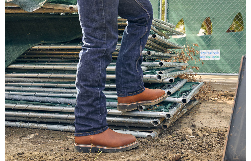 Close-up of a man, Nathan Menchaca, wearing brown Homeland work boots and dark jeans standing on a stack of metal construction fencing at a job site.